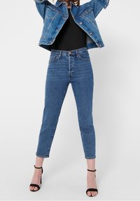Jeans in denim blu con vita alta, caviglie affusolate e chiusura a bottoni. Abbinati a una maglietta nera e a un giubbotto in denim azzurro chiaro.