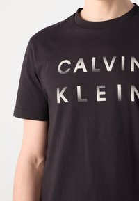 T-shirt en coton noir avec col rond, featuring grand texte blanc "CALVIN KLEIN" imprimé sur le devant dans un style gras et légèrement vieilli.