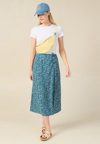 Keltainen kankainen vyölaukku, jossa on vetoketjullinen sulkija, käytössä valkoisen t-paidan päällä. Mallilla on päällä sininen kukkakuosiset kietaisuhame ja beigetan väriset sandaalit.