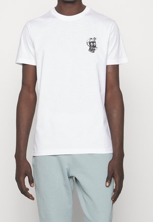 Homme portant un T-shirt blanc uni à manches courtes avec un petit motif noir de visage cartoon sur la partie supérieure gauche de la poitrine, associé à un pantalon bleu clair.