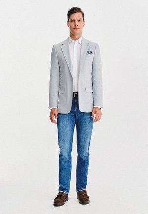 Mężczyzna stojący, ubrany w jasnoszary żakiet, białą koszulę, niebieskie dżinsy, brązowe buty oraz wzorzysty kieszonkowy chusteczkę.