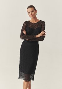 Rochie cocktail din dantelă neagră cu mâneci lungi, siluetă ajustată, tiv ondulat și modele florale complicate pe întreaga stofă.