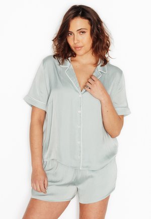Femme aux cheveux bruns mi-longs portant une chemise boutonnée manches courtes en satin bleu clair et un short assorti, posant devant un fond blanc.