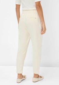 Lichtbeige taps toelopende broek met een elastische tailleband en twee achterzakken, gecombineerd met beige flatschoenen. Gladde, iets glanzende textuur.
