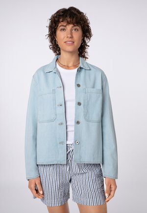 Frau mit lockigem Haar trägt eine hellblaue, geknöpfte Jacke über einem weißen Hemd und blau-weiß gestreifte Shorts, steht vor einem einfarbigen Hintergrund.