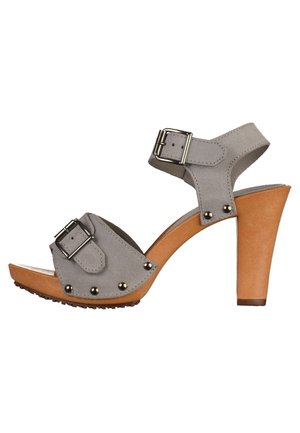 Szary zamszowy sandał na wysokim obcasie z dwoma regulowanymi klamrami, ozdobiony srebrnymi ćwiekami oraz obcasem przypominającym drewno z teksturowaną podeszwą.