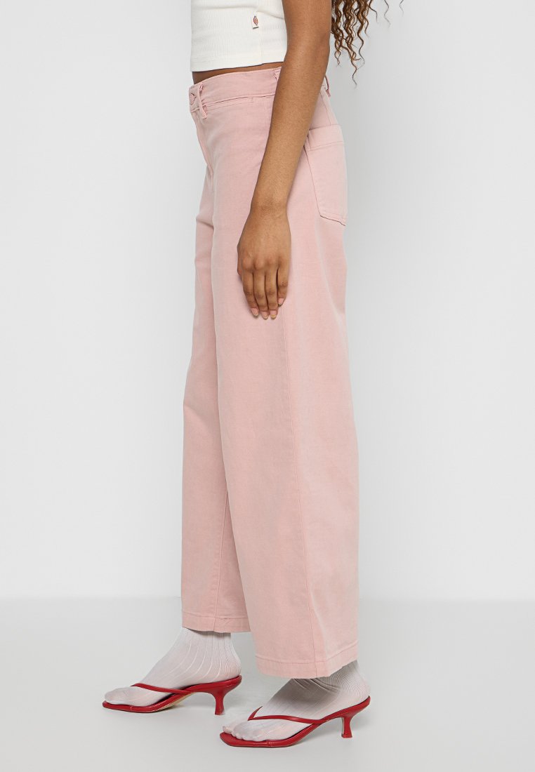 Jean large rose en denim, avec une taille haute, deux poches arrière et une coupe décontractée. Associé à des sandales à talons rouges et des chaussettes blanches.