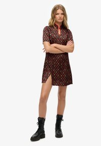 Kurzärmliges Kleid aus schwarzem Stoff mit rotem und gelbem Blumenmuster, Ausschnitt mit Schlitz und einem Seitenschlitz. Kombiniert mit schwarzen Kampfstiefeln.