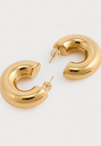 Złote kolczyki w kształcie okręgów o gładkim, zaokrąglonym designie. Każdy kolczyk posiada przekłuwaną bazę i przezroczyste zapięcie zapewniające pewne noszenie.