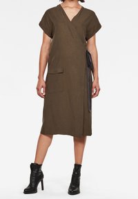 Vestido envolvente verde oliva hecho de lino, con mangas cortas, un bolsillo frontal y un detalle de lazo en la cintura; combinado con botas negras hasta el tobillo.