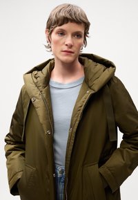 Olijfgroene pufferjack met een capuchon, voorzien van een rits aan de voorkant, zijzakken en verstelbare banden. In combinatie met een lichtblauw top.