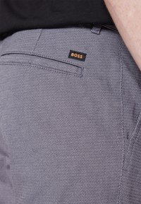 Šedé vzorované kalhoty s keprovou texturou. Mají černou cedulku s názvem značky "BOSS" v oranžové barvě. Viditelná je boční kapsa.