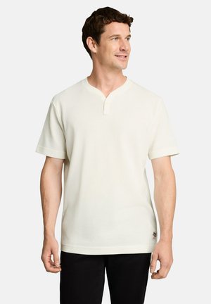 Man wearing a plain white short-sleeve shirt with a small V-notch collar, looking right, standing against a plain white background.