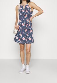 Robe sans manches bleu marine avec imprimé floral en rose et violet. Détail en forme de trou de serrure au niveau du cou, associée à des baskets montantes blanches.