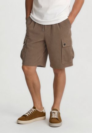 Bărbat purtând pantaloni scurți cargo de catifea maro și adidași maro cu tălpi albe, stând cu mâinile în buzunare pe un fundal simplu.