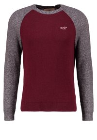 Maglione a collo rotondo con corpo bordeaux, maniche raglan grigie e piccolo logo di un uccello bianco sul lato sinistro del petto.