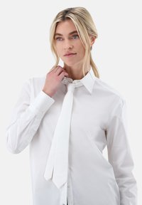 Camicia bianca con bottoni, collo a punta e maniche lunghe. Indossata con una cravatta bianca, il tessuto appare liscio e leggero.