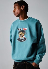 Sudadera azul-verde con cuello redondo, que presenta un diseño gráfico de un personaje de dibujos animados y el texto "GRMY" en el centro. Material de algodón.