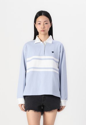 Hellblaues langärmliges Polohemd mit weißem Streifenmuster, weißem Kragen und kleinem dunkelblauen Herzemblem auf der Brust. Getragen mit schwarzen Shorts.