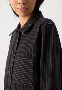 Czarna pasekowa kurtka z miękkiego materiału, z kołnierzykiem, zapięciem na guziki i kieszenią na piersi z pionowymi paskami.