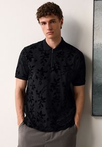 Fiatal férfi göndör hajjal, fekete rövid ujjú, virágmintás pólóingben és szürke nadrágban, zsebre dugott kézzel áll.