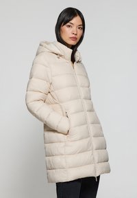 Abrigo acolchado beige claro con capucha, que cuenta con cierre con cremallera, bolsillos laterales y un patrón de acolchado horizontal para aislamiento. Textura suave, corte casual.