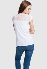 Camiseta blanca con un panel trasero de encaje floral, de manga corta y corte clásico, combinada con jeans ajustados azules.