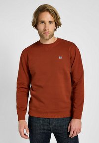 Felpa marrone a girocollo in cotone, con polsini e orlo ribattuti, e un piccolo logo blu Lee sul petto. Indossata con jeans scuri.