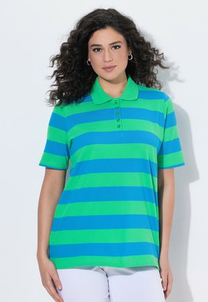 Kobieta o kręconych ciemnych włosach, ubrana w zielono-niebieską pasiastą koszulkę polo z krótkim rękawem i białe spodnie, stojąca na białym tle.