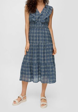 Vrouw die een mouwloze midi-jurk met V-hals en blauw patroon draagt, met een gelaagde rok en beige gevlochten sandalen met bandjes, staand tegen een effen achtergrond.