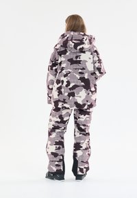 Kamoflage skidräkt i lila och grå nyanser, med huva, oversized passform och rutmönster. Tyget verkar tjockt och slitstarkt.