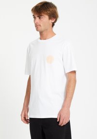 Valkoinen puuvillapaita lyhyillä hihoilla, jossa on pieni persikkakuvio vasemmassa rinnassa. Klassinen crew neck -muotoilu.