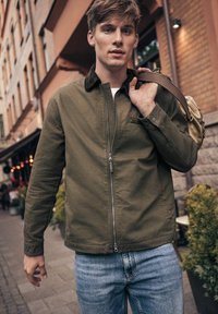 Oliven grønt zip-up jakke med brun krave, båret med lyseblå jeans; personen holder en tan taske over skulderen.