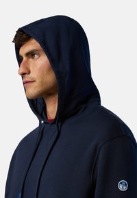 Marineblaue Kapuzenjacke mit Kordelzug, aus weichem Baumwollmaterial, gerippte Details an den Bündchen und einem runden Logo auf dem linken Ärmel.