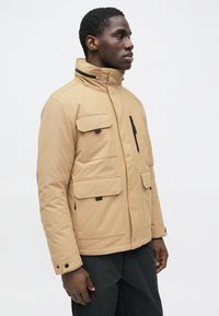 Mann, der nach rechts schaut, trägt eine beige Jacke mit mehreren Taschen und schwarze Hosen vor einem schlichten weißen Hintergrund.