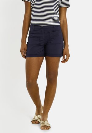 Femme portant un short bleu marine, un t-shirt rayé noir et blanc, et des sandales blanches à enfiler, debout devant un fond uni.