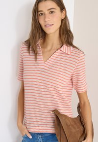 Jeune femme aux cheveux bruns portant un haut à manches courtes à rayures roses et un jean bleu, tenant un sac à main en daim marron à son côté.