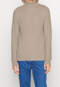 Beige gerippter Rollkragenpullover, getragen mit blauen Jeans, auf einer Person mit lockigem Haar, gezeigt von den Schultern bis zur Hüfte vor einem einfarbigen Hintergrund.