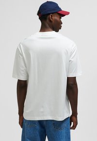 White short-sleeve t-shirt with a relaxed fit, featuring a smooth texture. The model wears blue denim pants and a navy and red cap.