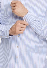 Vaaleansininen ja valkoinen raidallinen paita, pitkät hihat, nappikiinnitys messinkinupeilla. Rinnassa on tasku. Pehmeä puuvillakangas.