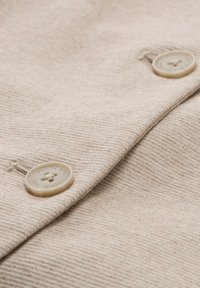 Tela beige con un patrón texturizado, que presenta dos botones de color claro con detalles de flores grabados, parcialmente ocultos debajo de una solapa.