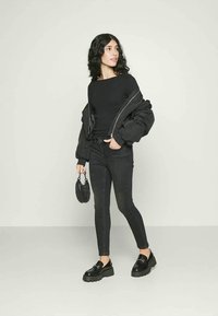 Μαύρο κοντό τζάκετ πάνω από ένα εφαρμοστό μαύρο μπλουζάκι μακρύ μανίκι, σε συνδυασμό με στενά μαύρα τζιν και στιλάτα μαύρα παπούτσια. Κρατάει μια μικρή μαύρη τσάντα.