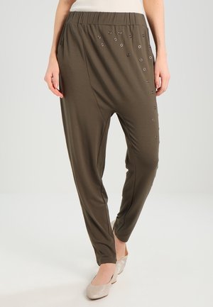 Kvinde iført olivengrønne haremsbukser med hulmønsterdetaljer på den ene side, kombineret med beige flade sko og en hvid top.