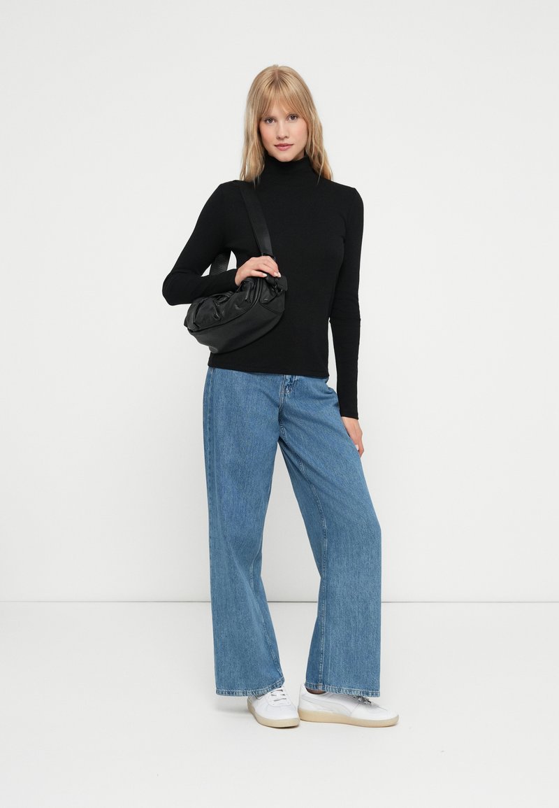 Maglione nero a collo alto, jeans azzurri a gamba larga e borsa nera a tracolla. La modella indossa scarpe da ginnastica bianche, in piedi su uno sfondo bianco.