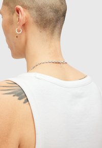 Witte geribbelde tanktop met een ronde halslijn, gecombineerd met een zilveren ketting. Een tatoeage van zwarte veren is zichtbaar op de schouder.