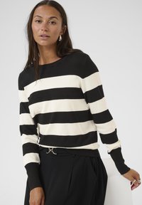 Pull rayé noir et crème, avec des poignets côtelés et un col rond, associé à un pantalon noir taille haute et une ceinture avec des détails en métal argenté.