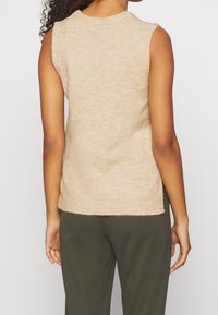 Persona che indossa un top a maglia beige senza maniche e pantaloni verde oliva scuro, vista da dietro su uno sfondo neutro.