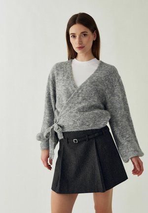 Femme portant un pull gris croisé noué à la taille, un haut blanc et une jupe plissée foncée avec une ceinture, debout devant un fond uni.
