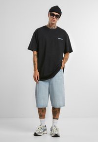 Fekete oversized póló logóval, világoskék farmer rövidnadrág és szürke sportcipő. Kiegészítők közé tartozik egy fekete sapka és napszemüveg.