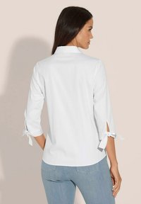 Witte blouse met driekwart mouwen, voorzien van zijslits die vastgebonden zijn met strikjes. Gladde textuur en een klassiek kraagontwerp, gecombineerd met lichtblauwe jeans.